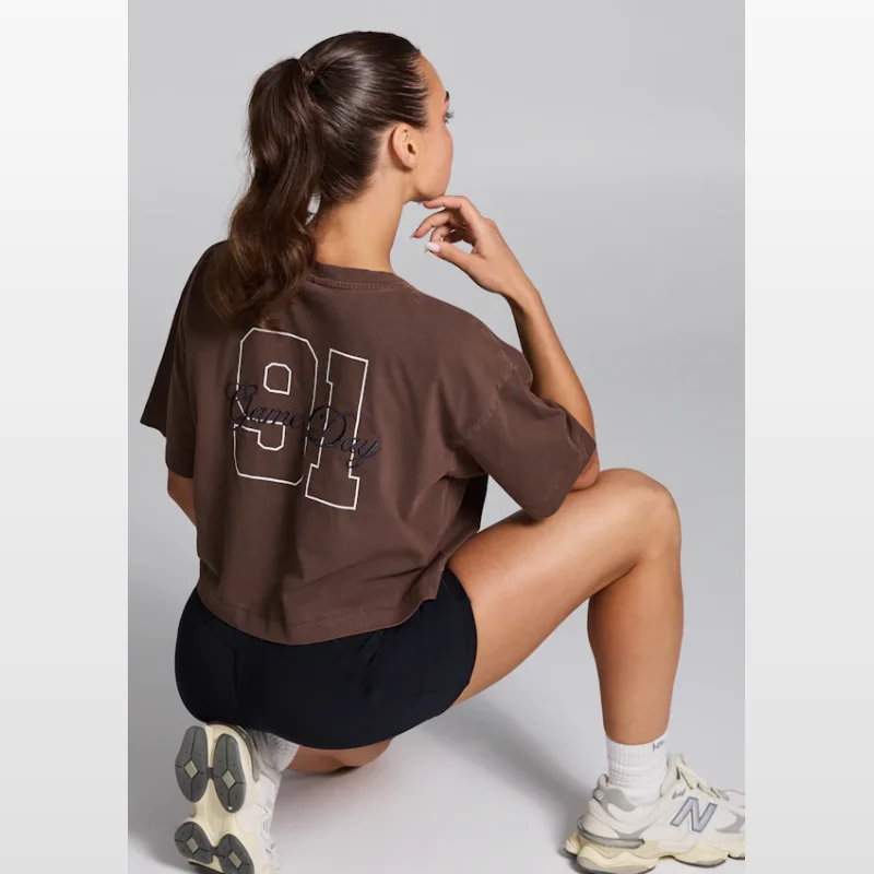 Woman wearing a brown Rockwear V-neck hip length t-shirt with a 'Game Day 91' graphic on the back, paired with black bike shorts and sneakers.