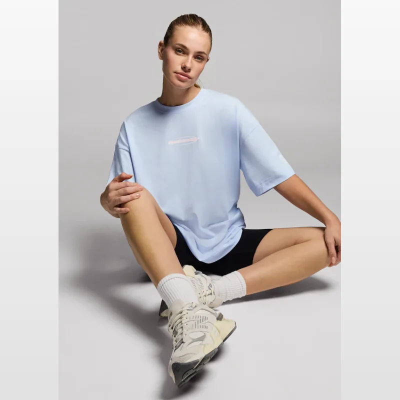 Woman wearing a light blue Rockwear baggy fit cotton t-shirt with a small pink logo detail on the chest, black bike shorts, white socks, and cream sneakers, sitting cross-legged against a grey background.