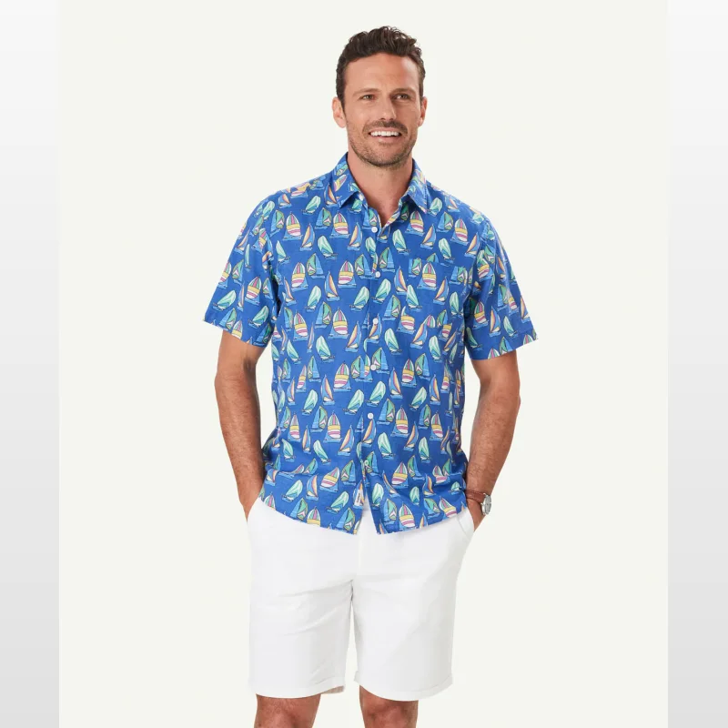 Man wearing a royal blue short sleeve shirt with a repeating print of colourful sailboats. He is also wearing white shorts.