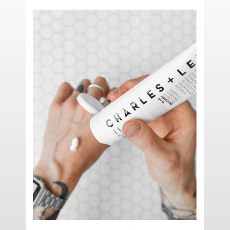 A hand squeezing a white tube of Charles + Lee SPF 15 Moisturiser onto another hand, with a small amount of cream visible on the palm.