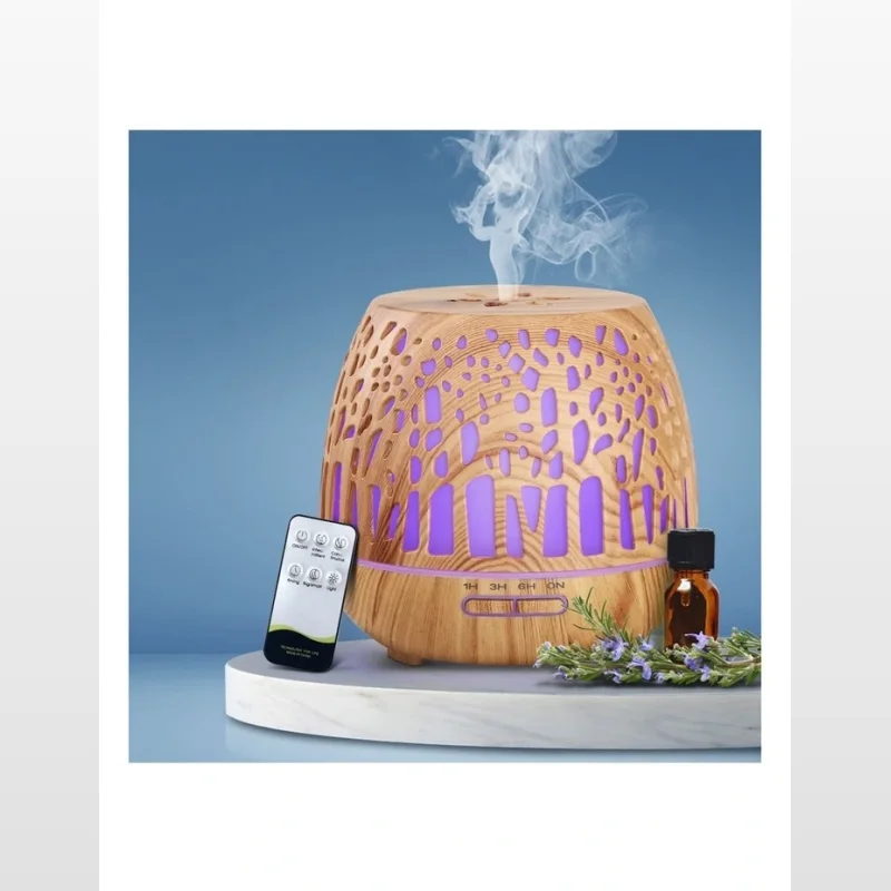A wood grain finish ultrasonic aroma diffuser with purple LED lights, emitting a stream of vapour, with a remote control and a small bottle of essential oil next to it.