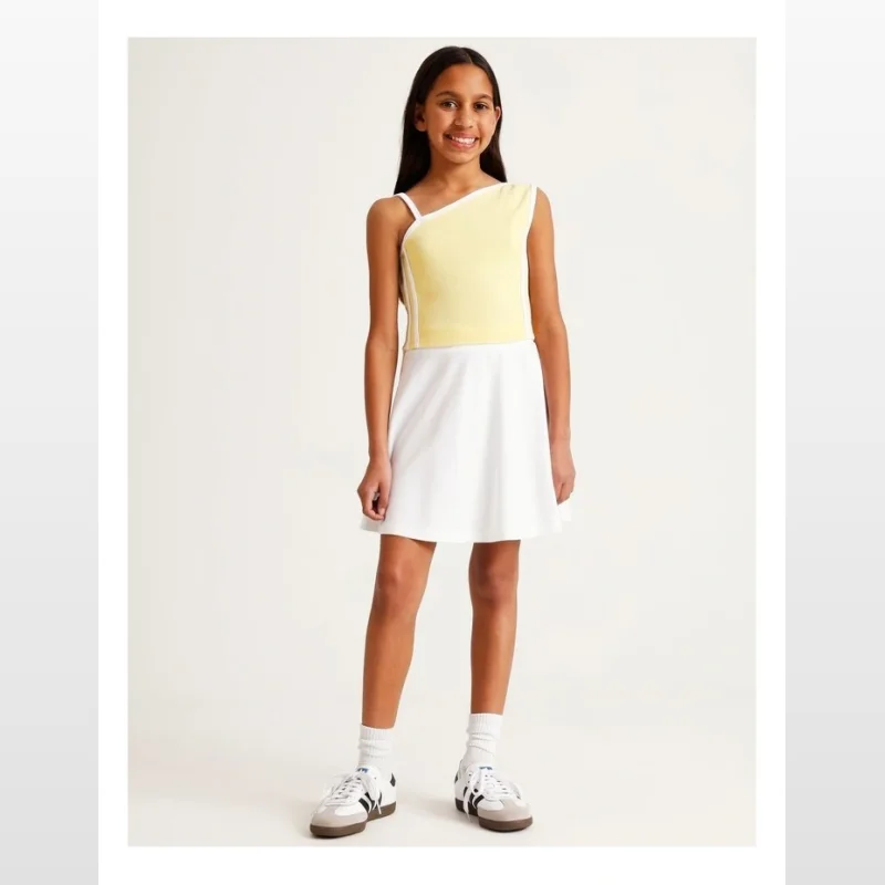 A young girl wearing a yellow one-shoulder ribbed top with white trim and a white skirt, standing against a white background.