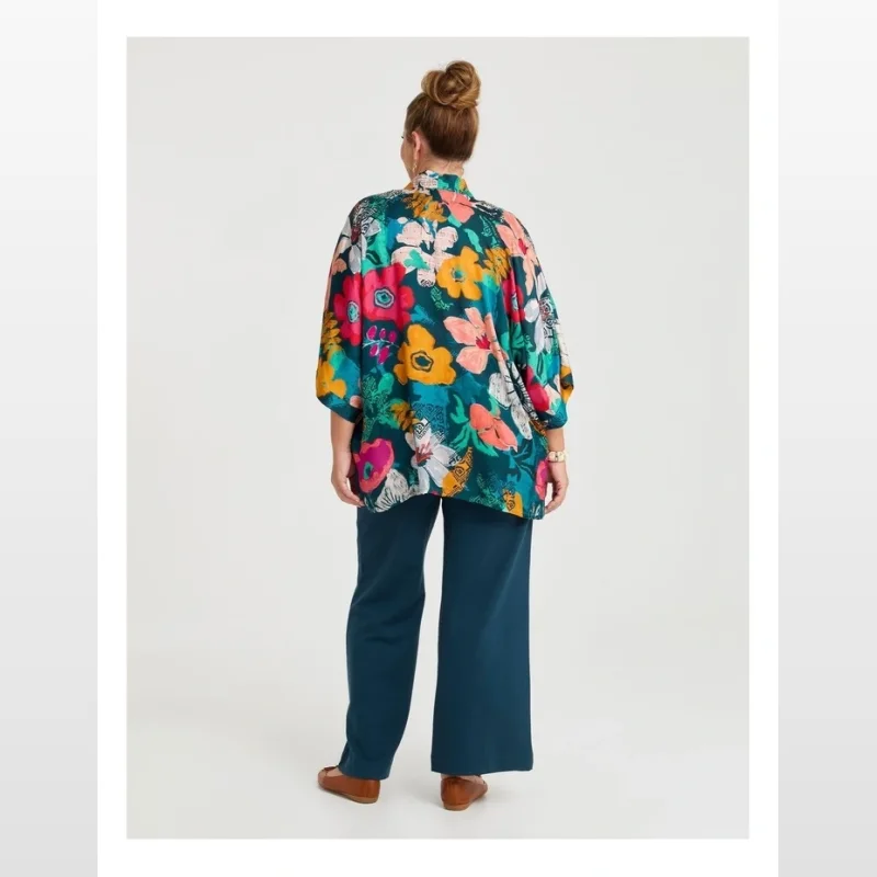 Woman wearing a dark teal kimono with a vibrant floral print featuring pink, orange, and white flowers, and 3/4 length sleeves. She is also wearing wide-leg teal trousers.