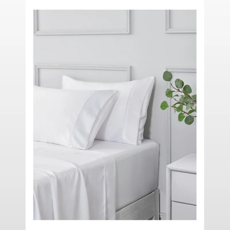 A close-up shot of a white matte satin sheet set on a bed, featuring two pillowcases and a fitted sheet.