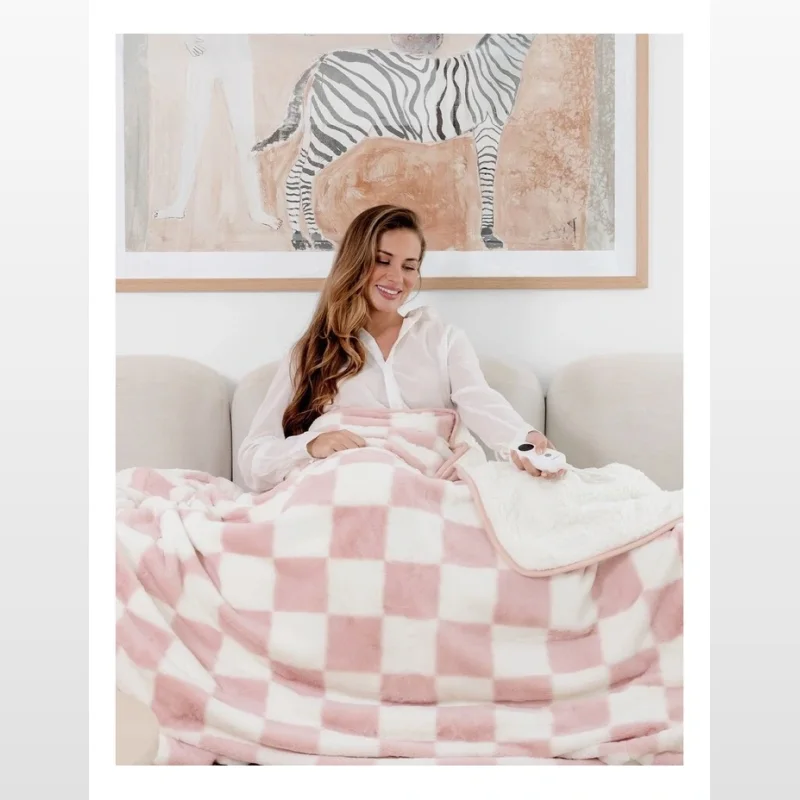 Woman sitting on a couch wrapped in a pink and cream checkered plush electric heated throw blanket.
