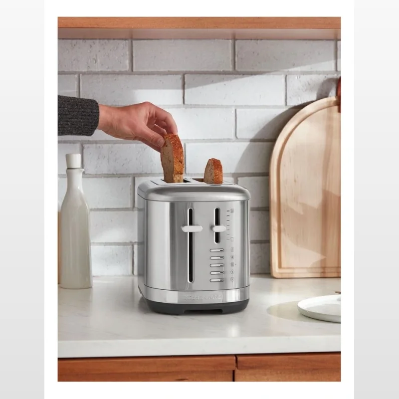 A person is placing a slice of bread into a silver KitchenAid 2-slice toaster on a marble countertop.