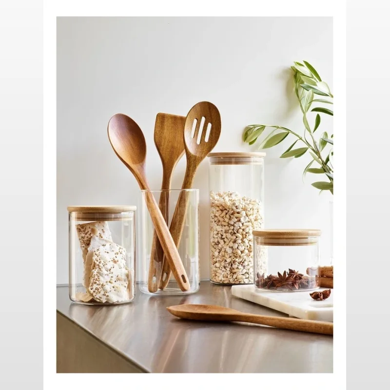 A collection of glass storage canisters with bamboo lids, one filled with popcorn, another with star anise, and one with biscuits. Wooden utensils are also displayed.