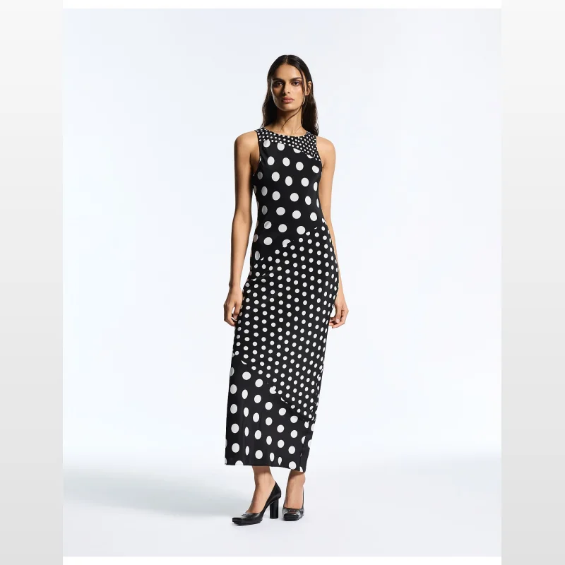 Woman wearing a sleeveless, high-neck, black and white polka dot maxi dress with a bias-cut silhouette.