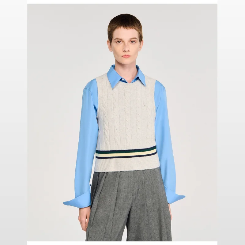 Woman wearing a cream-coloured sleeveless cropped cable knit Sandro Paris jumper vest with contrasting navy, green, and yellow stripes around the hem, layered over a light blue collared shirt.
