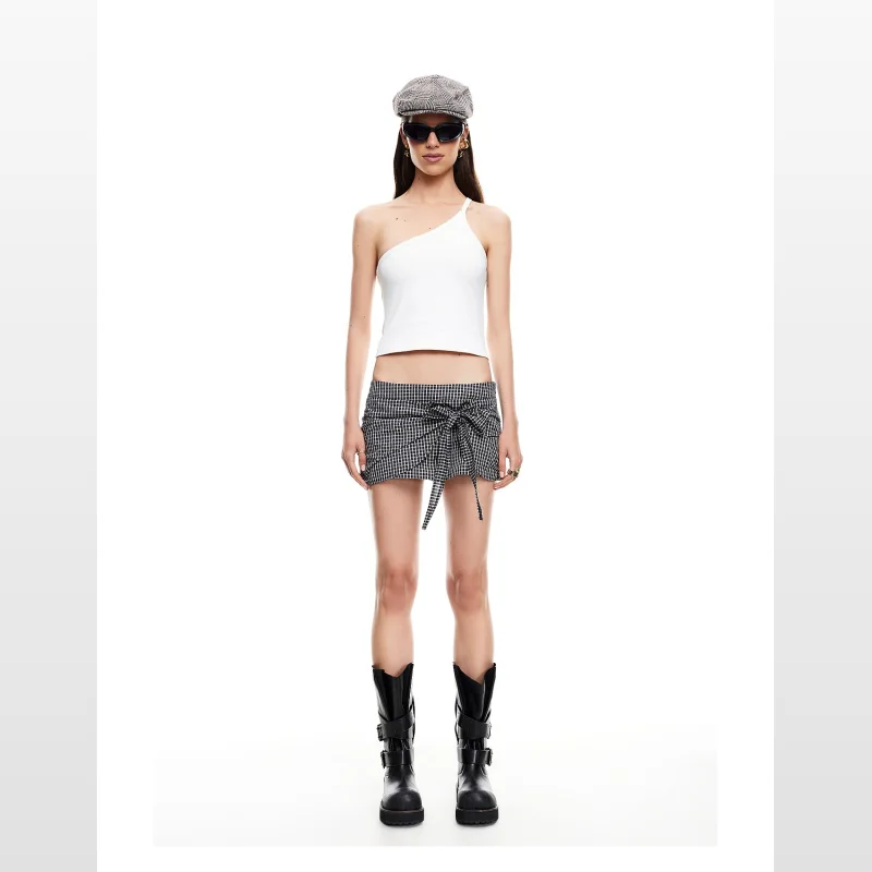 Woman wearing a white one-shoulder top and a black and white gingham mini skort with a tie front detail, black sunglasses, a grey flat cap, and black buckle boots.