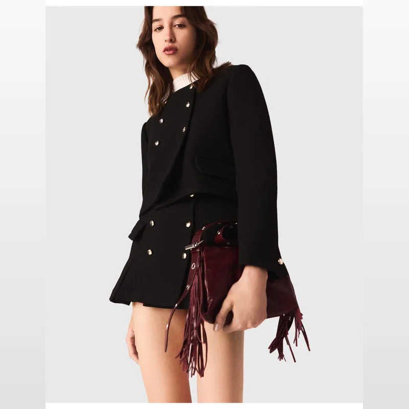 Woman wearing a black Maje Vabatte cropped blazer with gold double-breasted buttons and a round collar, holding a burgundy fringed handbag.