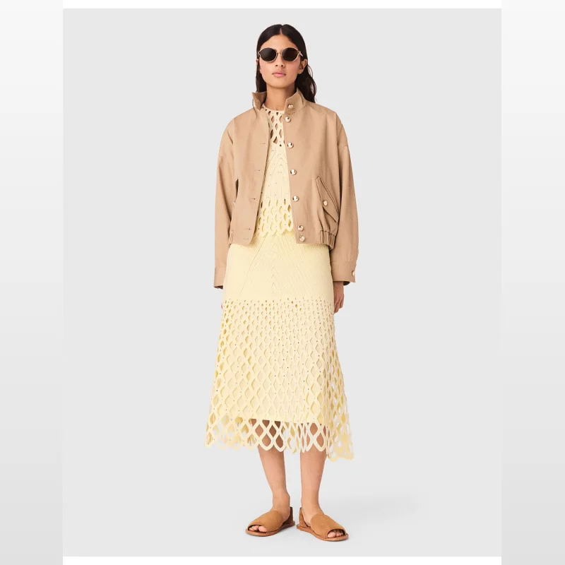 Woman wearing a long, pale yellow crochet midi skirt with a diamond pattern, silver rhinestones, and a scalloped hem, paired with a beige bomber jacket and tan sandals.