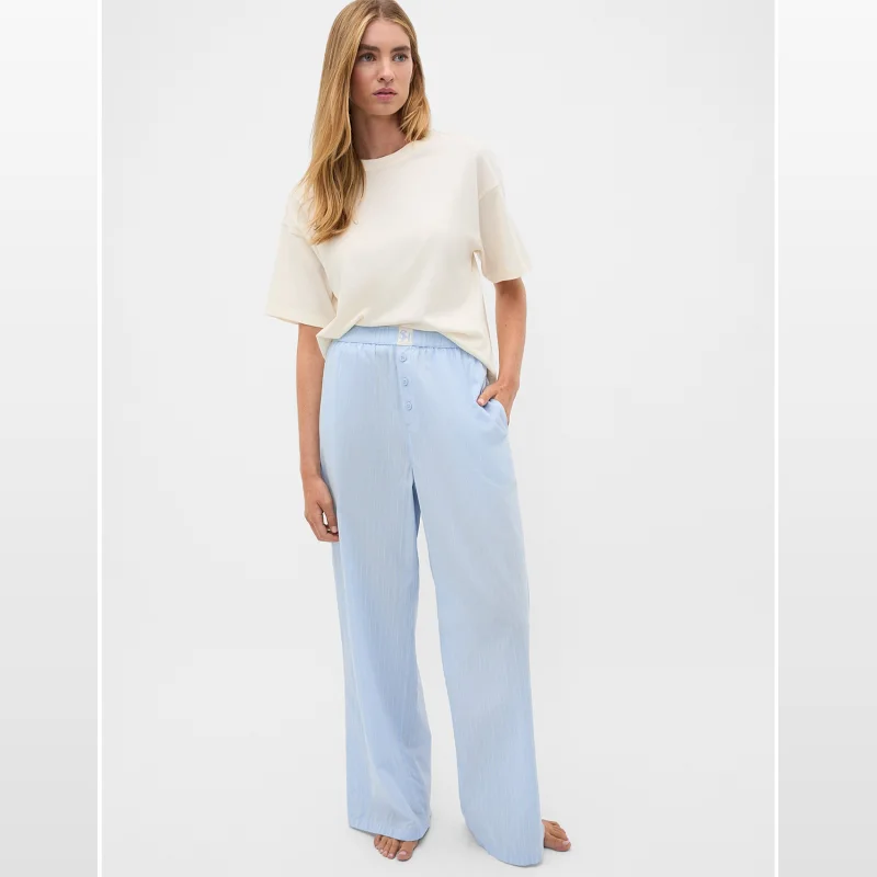 Woman wearing a cream t-shirt and light blue pinstripe pyjama pants with an elasticated waistband and button-up detail.