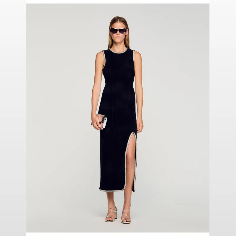 Woman wearing a black sleeveless Sandro Paris Guoda velvet knit maxi dress with a boat neck, rhinestone trim, and a high front slit, paired with silver strappy heels and sunglasses.