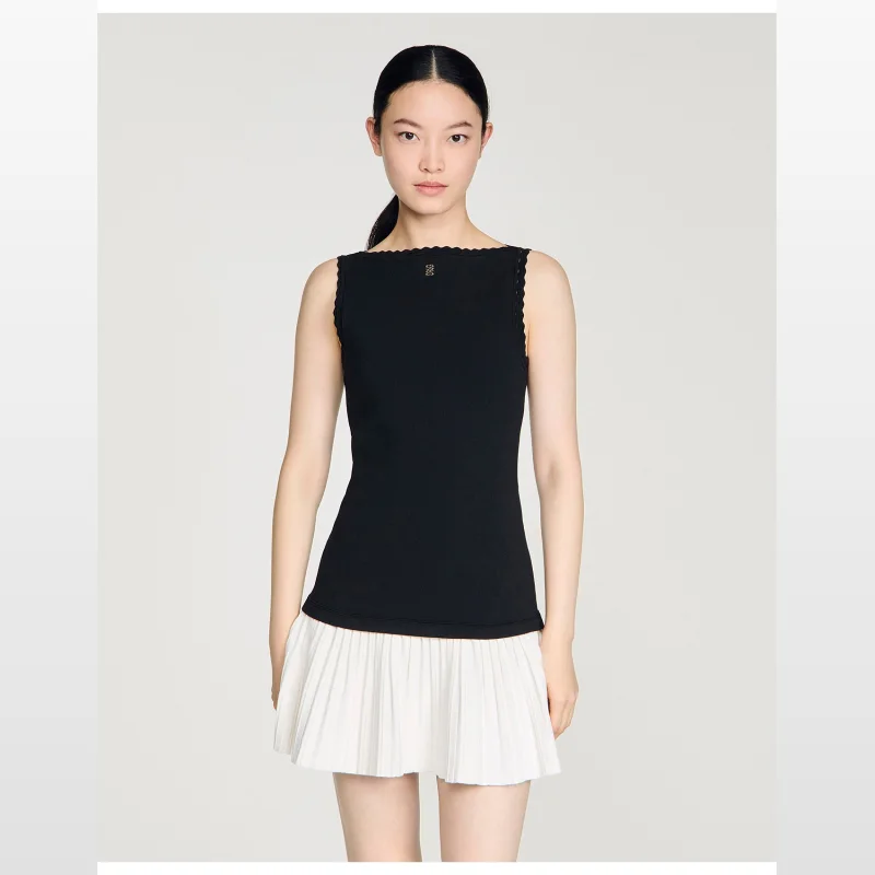 Woman wearing a black sleeveless Sandro Paris knit top with a white pleated mini skirt. The top has a boat neckline with a scalloped edge and a small gold logo detail.