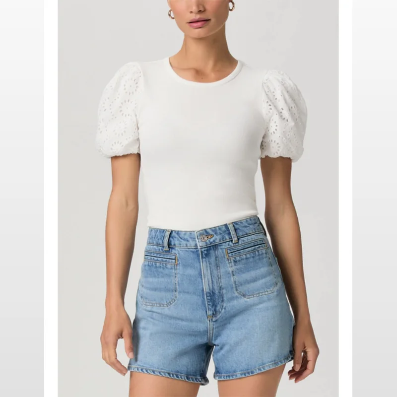 Model wearing PAIGE Dani high-rise, light wash denim shorts with a finished hem and double welt stitch pockets, paired with a white ribbed top featuring puff sleeves with eyelet embroidery.