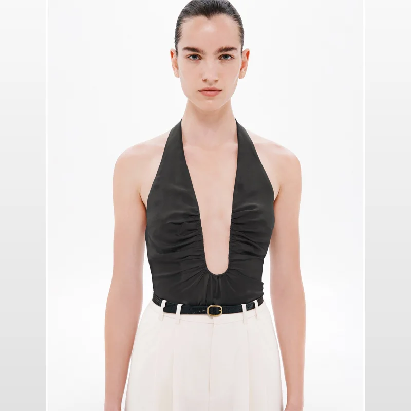 Model wearing a black MISHA 'Alusia' silk halter top with a plunging neckline and ruched detailing, paired with cream trousers and a black belt.