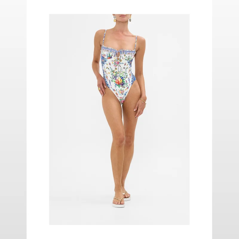 Woman wearing a white one-piece swimsuit with a vibrant floral and tile print, featuring ruched cups, a tie front, and adjustable straps.