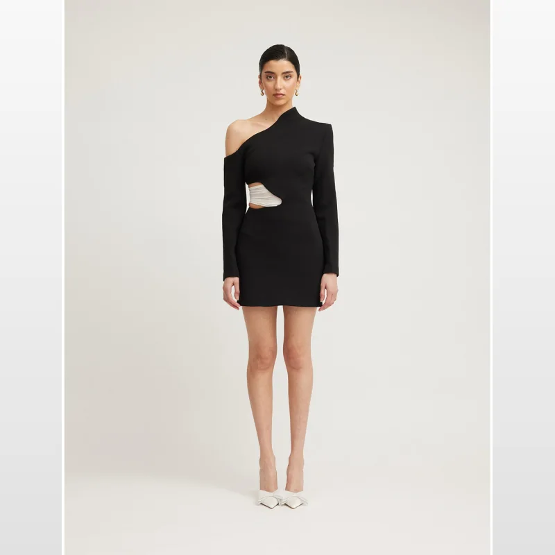 Model wearing the TOJHA Martina One Shoulder Cut Out Mini Dress in black, featuring a single long sleeve, asymmetric neckline, and a curved waist cut out with white mesh detail.