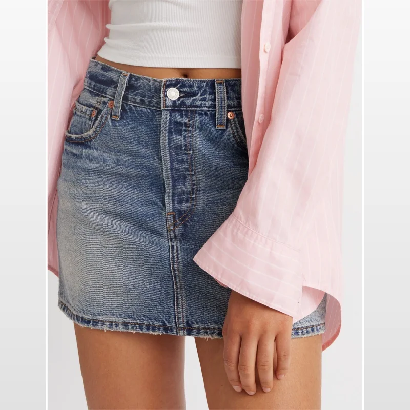 Close-up of a woman wearing a blue denim Levi's icon skirt with a mid-rise waist and button fly, paired with a white ribbed tank top and a pink striped shirt.