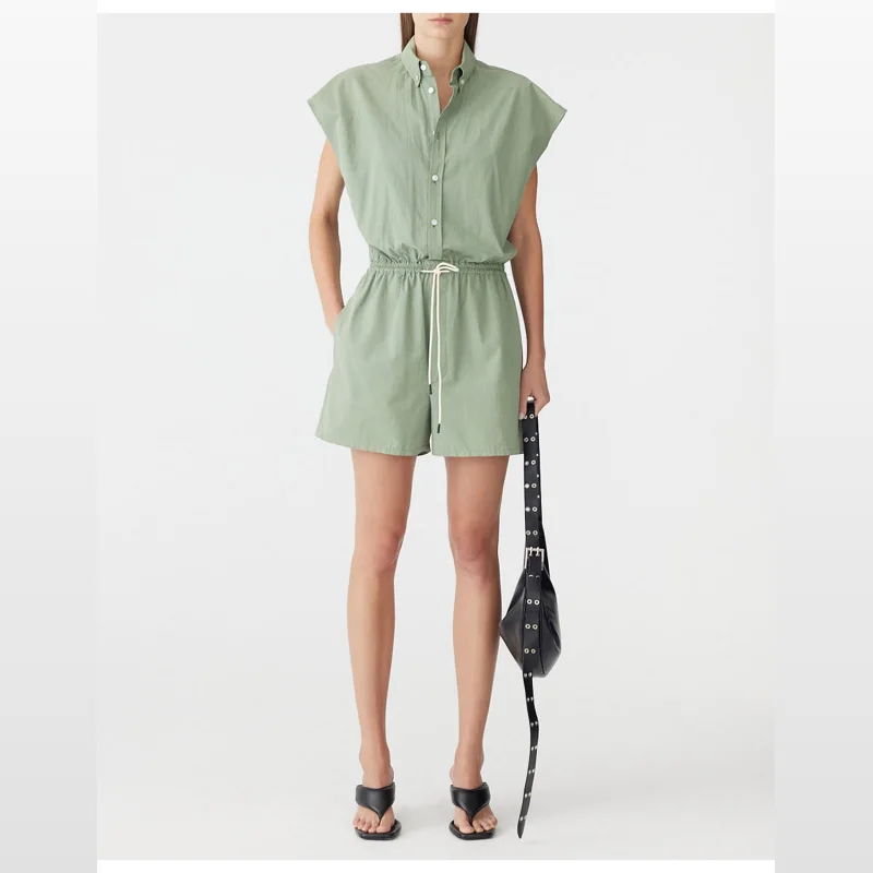 Woman wearing a sage green, short-sleeved, collared mini jumpsuit with an elasticated drawstring waist and white drawstrings. She is holding a black studded shoulder bag and wearing black thong sandals.