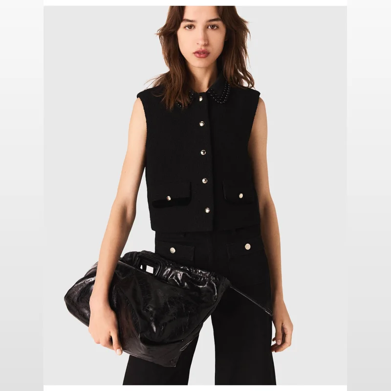 Woman wearing a black Maje LOBYNETTE TOP sleeveless crop vest with a pearl-embellished collar and gold button details, holding a black patent leather clutch.