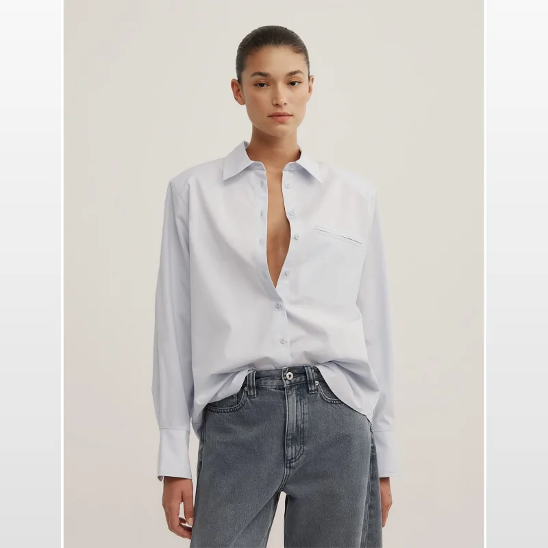 Woman wearing a light blue long-sleeved collared shirt with subtle shoulder pads and a button-through placket, paired with grey denim jeans.