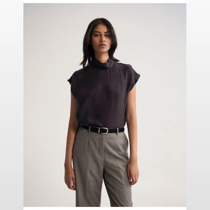 Woman wearing the OXFORD Shelly Cupro Top in charcoal grey, featuring a high neckline and cap sleeves, paired with grey patterned trousers and a black belt.