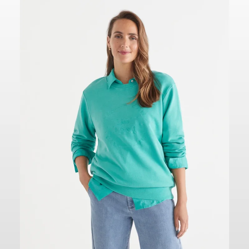 Woman wearing a teal Sussan 'This Goes With That' Heritage Sweat with embroidered crest and 'EST 1981' detail, layered over a collared shirt and paired with light wash jeans.