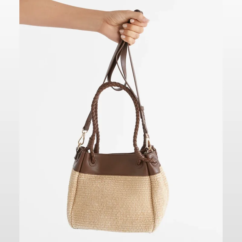 A hand holds a Sussan Spliced Raffia Bucket Bag, featuring a woven raffia body and brown leather accents.