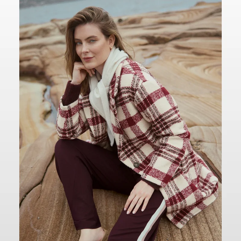 Woman wearing a red and cream check single-breasted coat, cream scarf, and burgundy trousers, sitting on rocks by the sea.