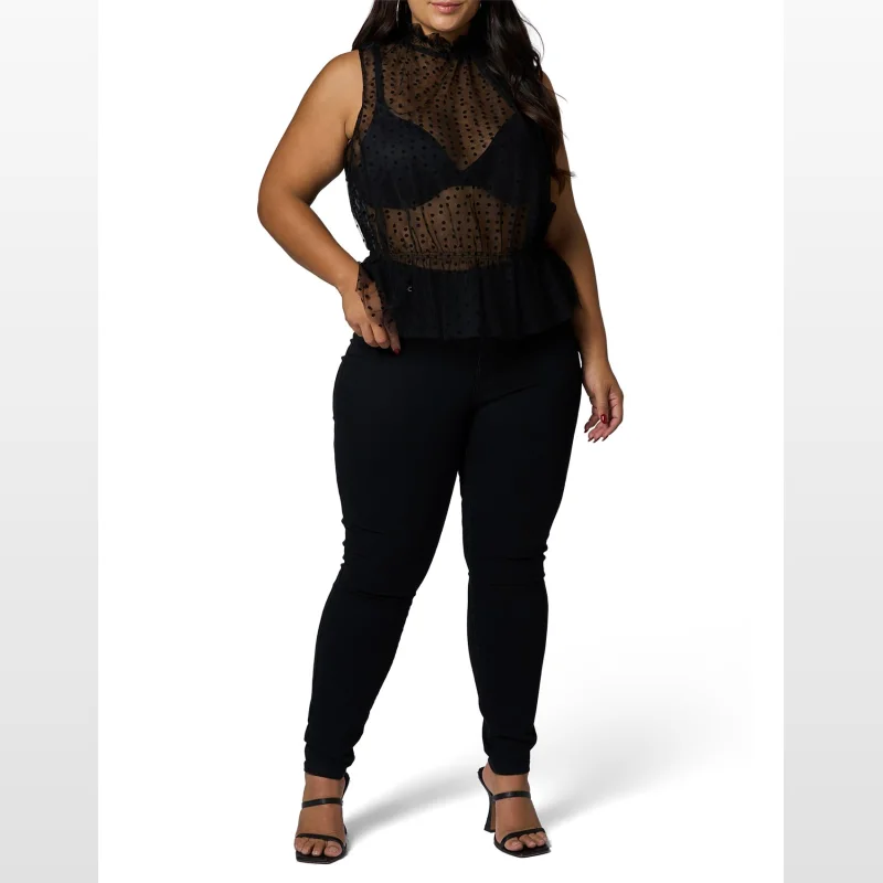 Woman wearing a black sheer polka dot mesh top with a high frill neck and peplum hem, paired with black skinny jeans and black heels.