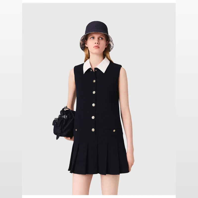 Woman wearing a navy blue sleeveless Maje Rondine tweed knit mini dress with a white collar and gold buttons, holding a black handbag and wearing a navy bucket hat.