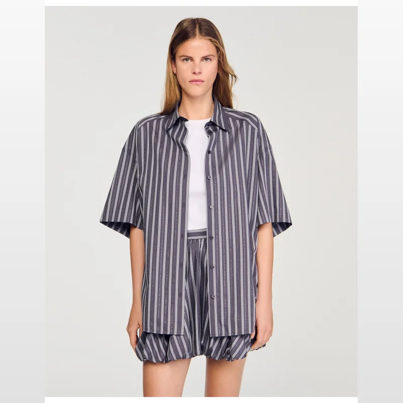 Woman wearing a Sandro Paris oversized grey and white striped short-sleeve shirt with rhinestone detailing, featuring a shirt collar and button fastening.