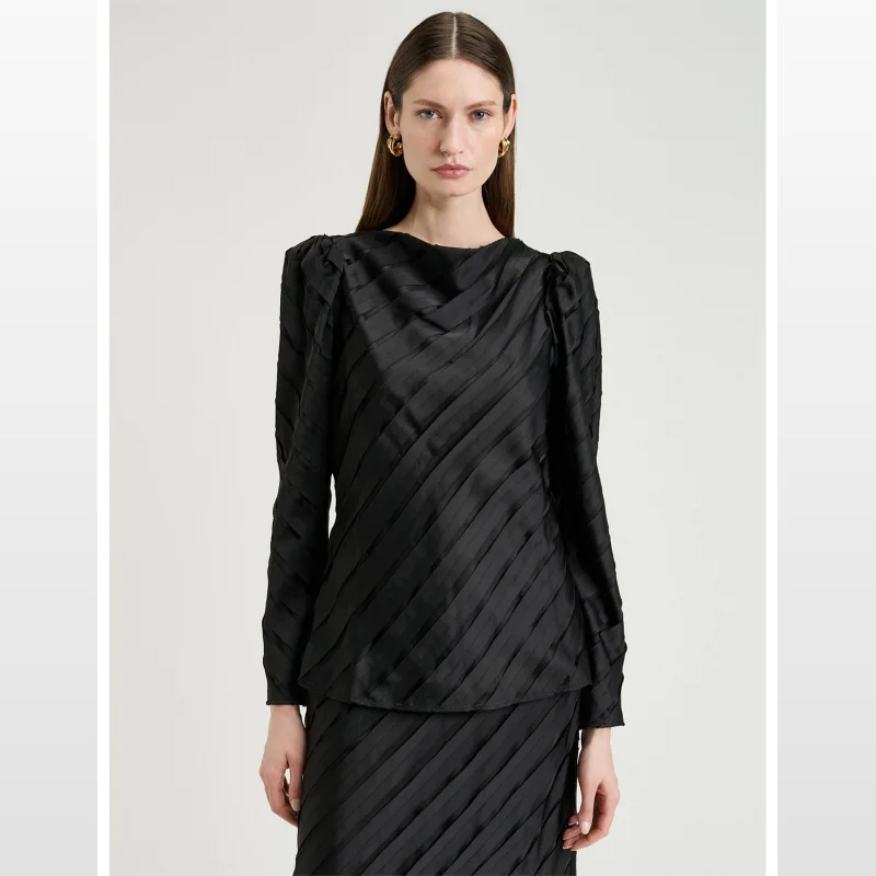 Woman wearing a black long-sleeved Veronika Maine blouse with a bias-cut, draped silhouette and diagonal pleating. The blouse has a smooth back and is hip length.