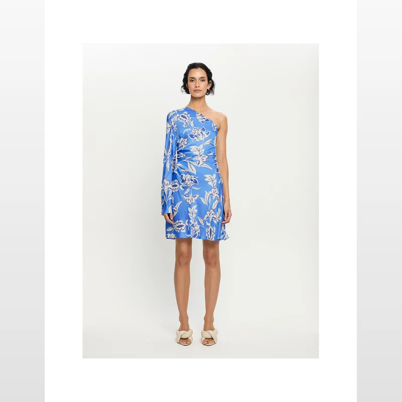 Woman wearing a blue KIVARI Ren One Sleeve Mini Dress with a white floral print, asymmetrical neckline, and a single flared sleeve.