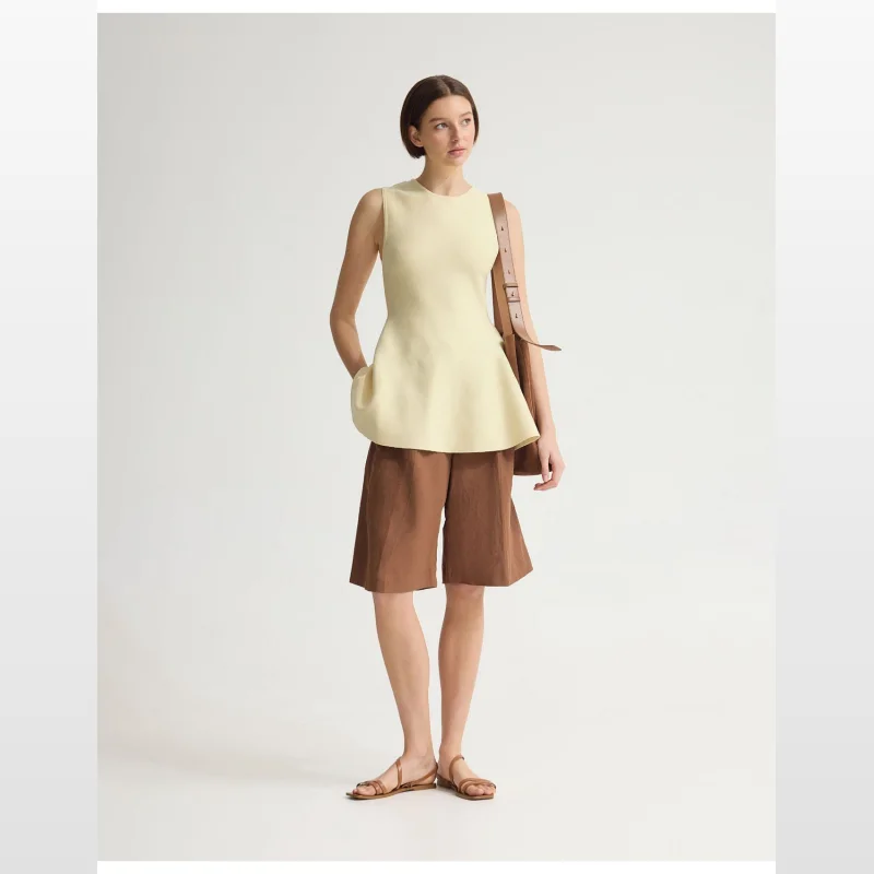 Model wearing a pale yellow sleeveless knitted peplum top with brown shorts and tan sandals, carrying a brown shoulder bag.