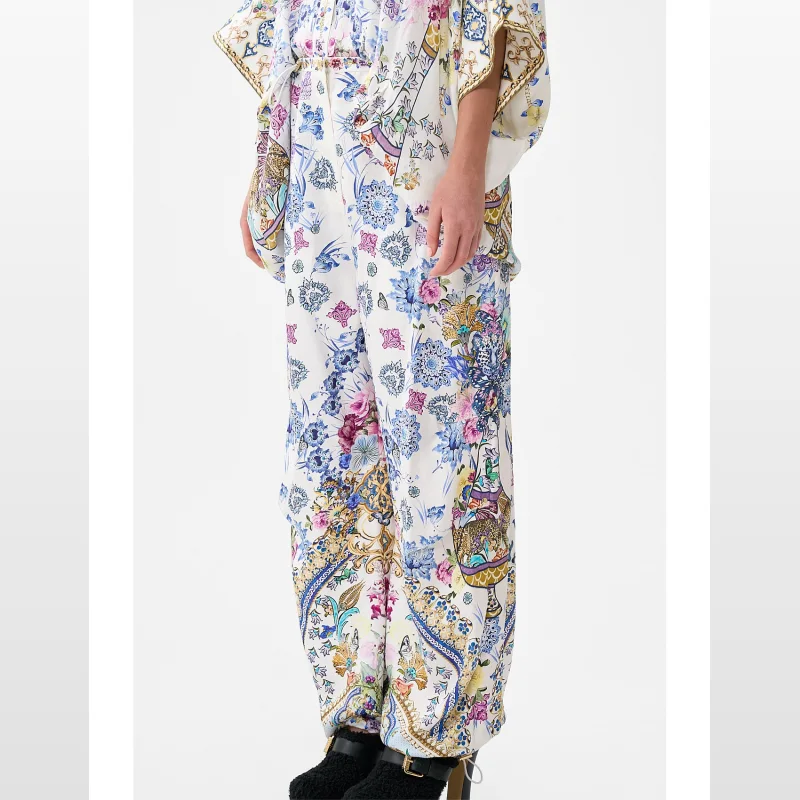 Close-up of a person wearing CAMILLA All About That Lace Parachute Pants with a vibrant, intricate floral and ceramic-inspired print on a white background.