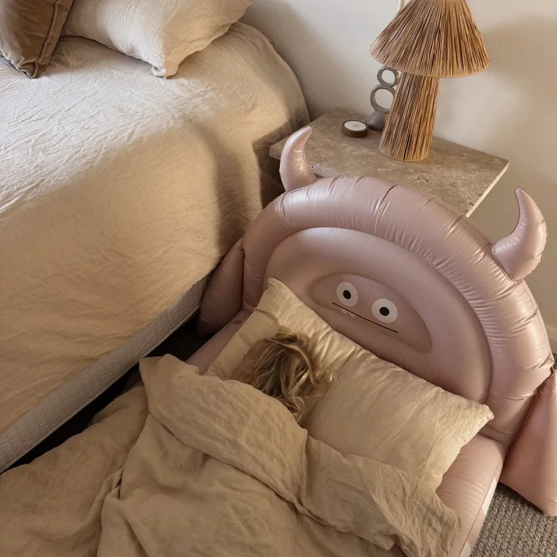 A child sleeps on a pink inflatable monster-shaped air mattress with horns and a cute face, next to a larger bed.