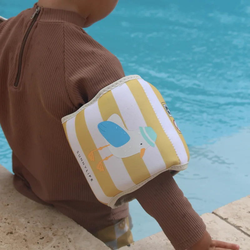 Close-up of a child wearing yellow and white striped Sammy the Seagull floaties on their arm, with a swimming pool in the background.