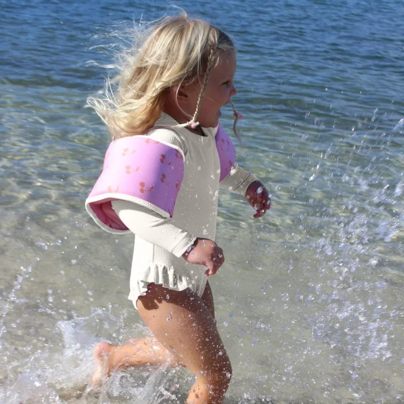 A young girl with blonde hair running through shallow water at the beach, wearing a white long-sleeved swimsuit and pink floaties with a cherry pattern on her arms.