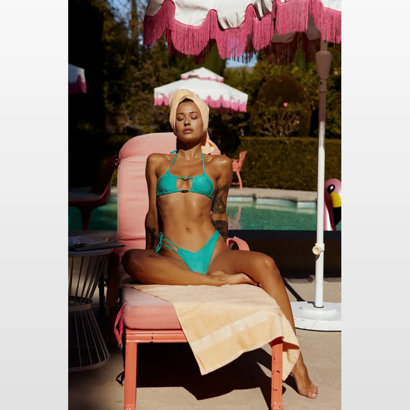 Woman wearing a green scoop neck halterneck bikini top with adjustable tie-up halterneck and tie back with gold toggles, sitting on a pink sun lounger by a pool.