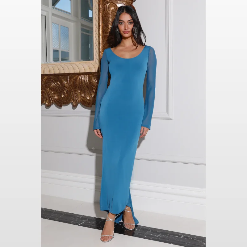 Woman wearing a long-sleeved blue maxi dress with a scoop neckline and a tie-up back, standing in a room with ornate mirror and marble flooring.