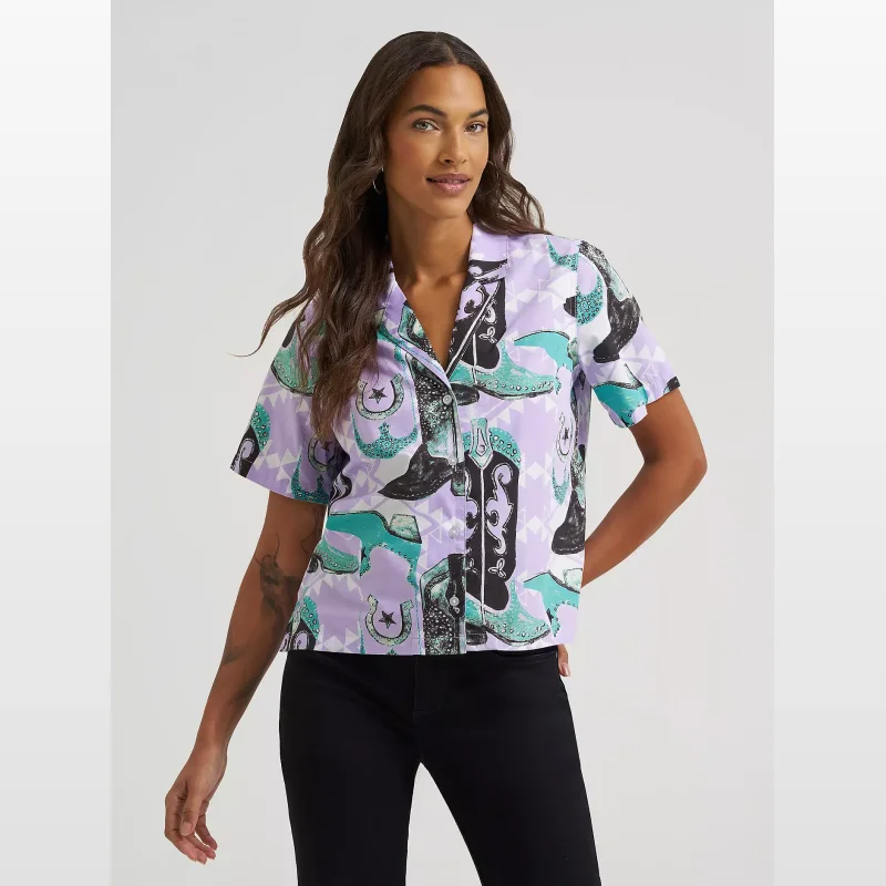 Woman wearing a short-sleeved, collared Wrangler camp shirt with a bold print of cowboy boots and horseshoes on a lavender background.