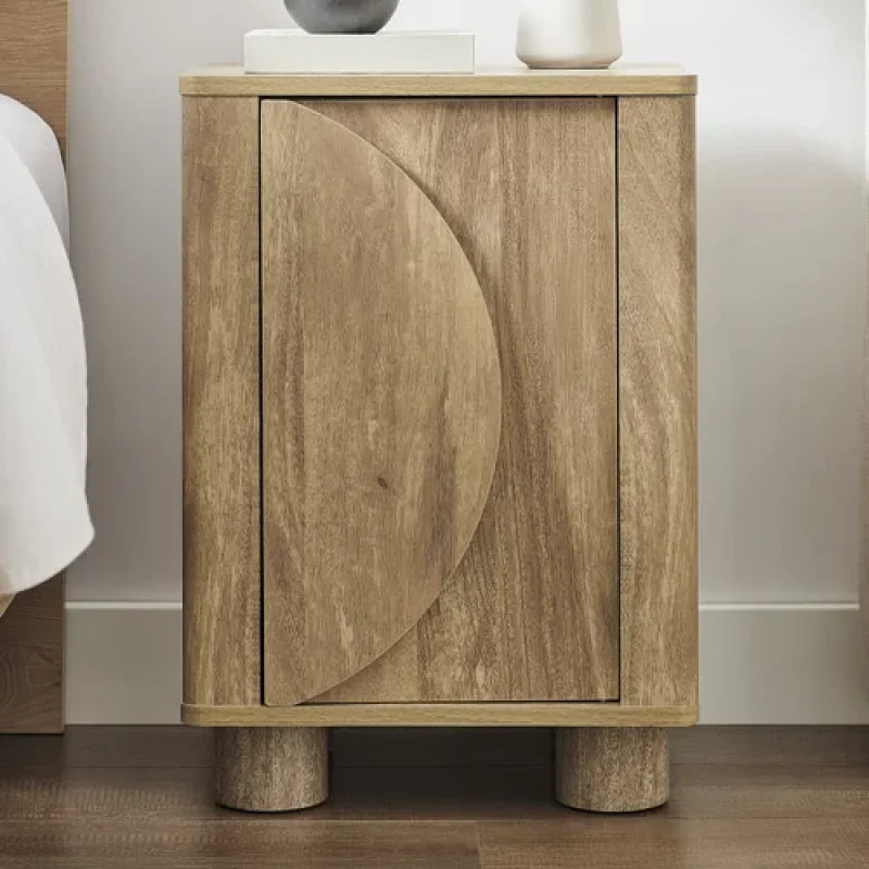 A wooden bedside table with a curved door detail and cylindrical legs, placed next to a bed.