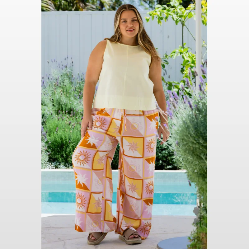 A plus-size woman wearing a pale yellow sleeveless top and wide-leg trousers with a colourful sun and square print, standing outdoors by a pool.