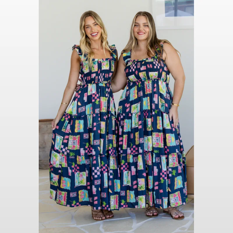 Two women wearing the Orangesherbet Darcey Maxi Dress in Tuscan Sun Print. The dress features a navy blue base with a colourful print of buildings, palm trees, and checkered patterns. It has a smocked bodice, square neckline, and tiered skirt with ruffle