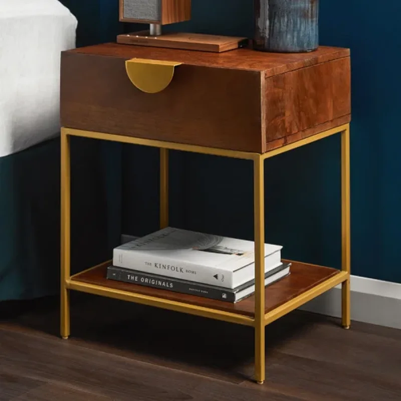 A dark brown mango wood bedside table with a gold semi-circle drawer handle and a lower display shelf supported by thin gold metal legs.
