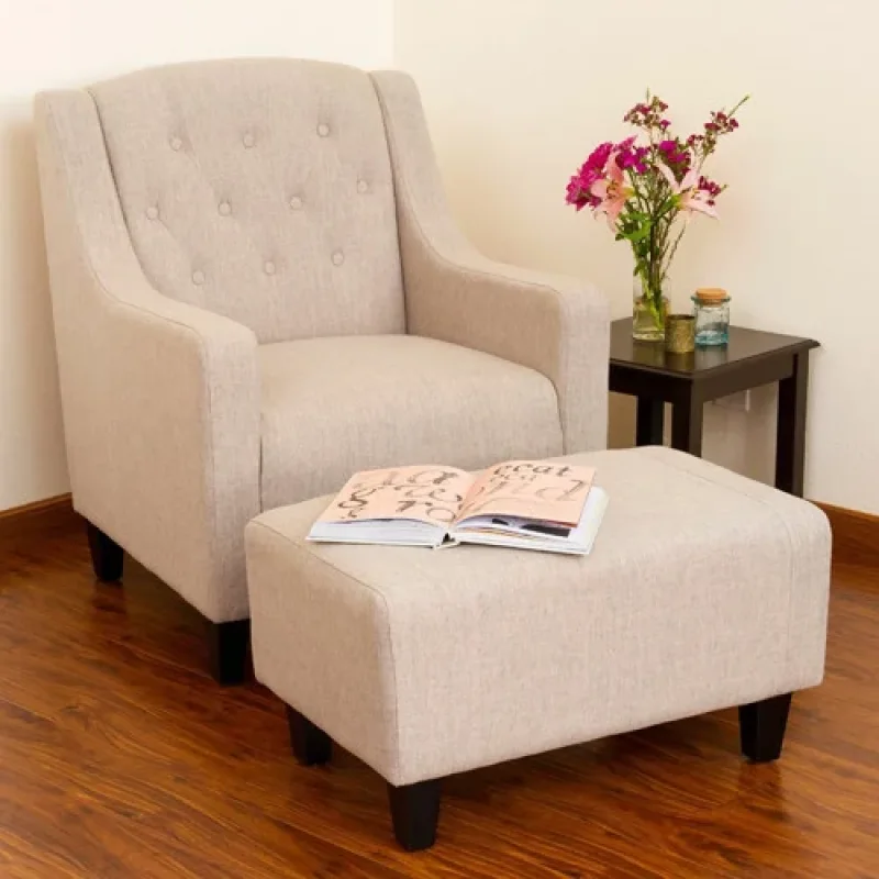 A beige fabric armchair with a matching footstool in front of it, with a book open on the footstool and a small side table with flowers behind it.
