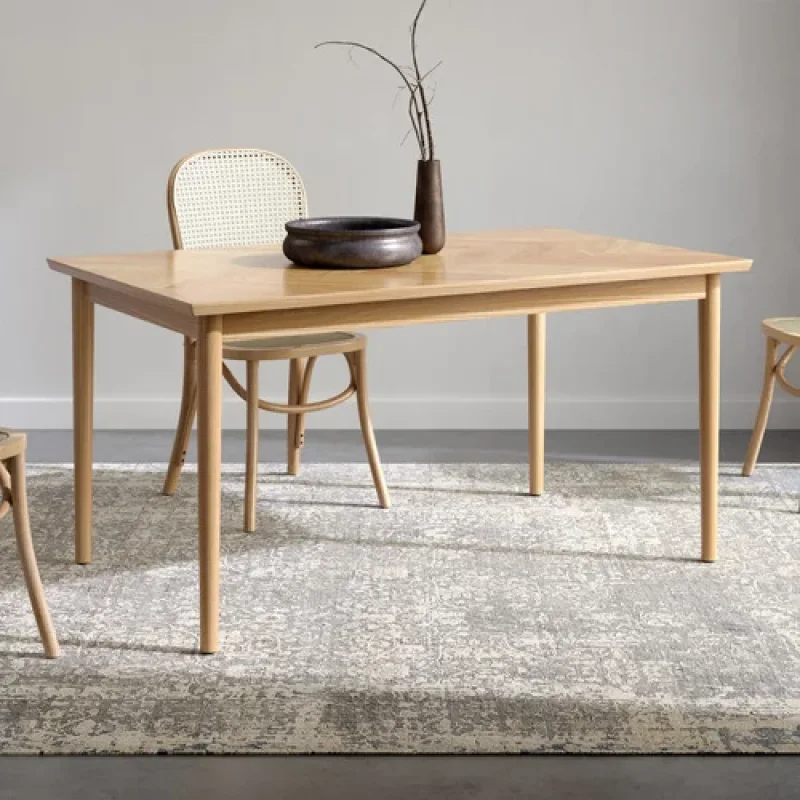 A light oak wood dining table with a herringbone pattern on the tabletop and slender tapered legs, shown with cane-backed chairs and a decorative bowl and vase.
